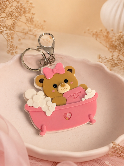 A cute bear-shaped keychain in a pink bathtub with white bubbles and a pink bow, resting on a white dish with dried flowers in the background.