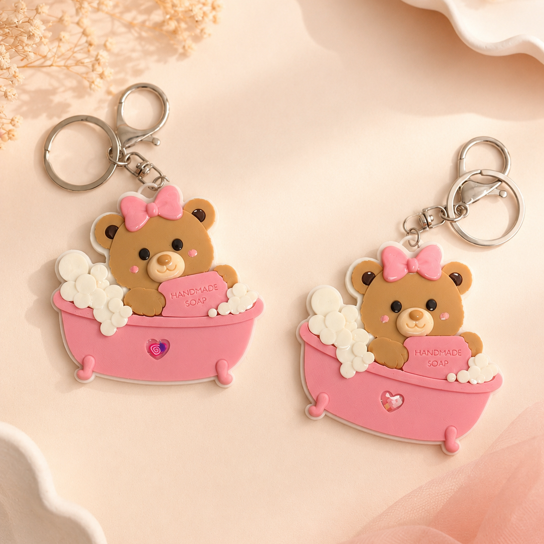 Two matching keychains featuring a cute brown teddy bear sitting in a pink bathtub with white bubbles and a pink bow, displayed on a soft beige surface with dried flowers and decorative props.