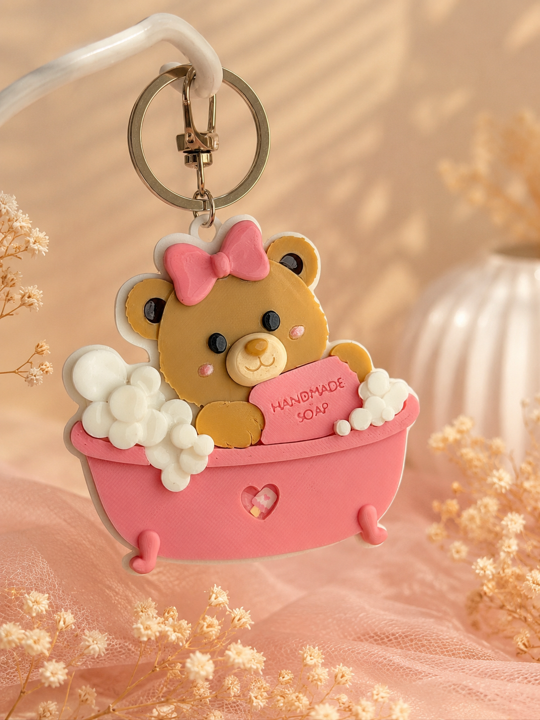 A cute bath-themed keychain charm featuring a yellow teddy bear sitting in a pink bathtub with white bubbles and a pink soap bar that reads 'HANDMADE 2023'. The keychain is shown hanging against a soft peach background with decorative dried flowers.